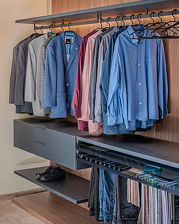 Offener Kleiderschrank: Vor einer Holzwand befinden sich Regale, mit Kleiderstangen an denen Hemden und Hosen aufgehängt sind. Die Regale sind von hinten beleuchtet.