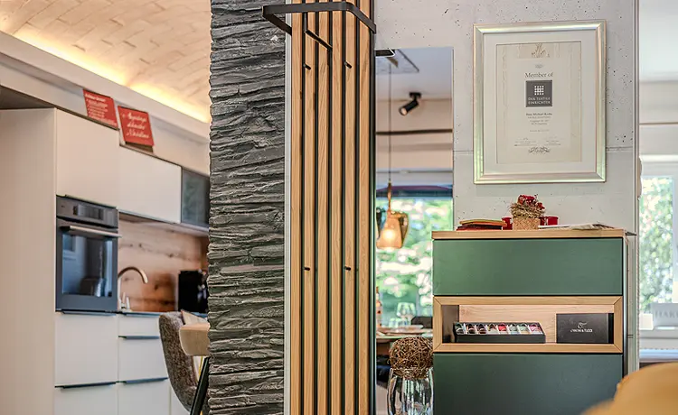 Wohnbeispiel Garderobe: Blick auf eine Garderobenleiste aus Holzlamellen. Daneben befindet sich ein Sideboard mit grüner Front und einem beleuchteten Fach aus hellem Holz. Im Hinergrund ist die Küche zu sehen.