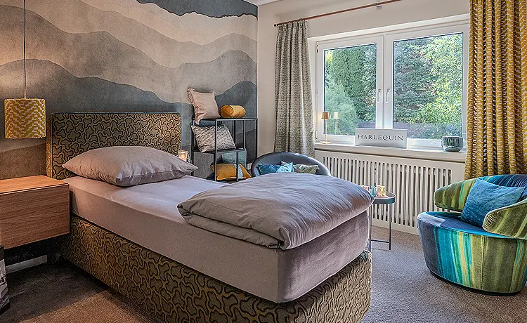 Wohnbeispiel Schlafzimmer: Auf der linken Seite befindet sich ein Bett, dahinter befindet sich ein Fenster.Zwischen Bett und Fenster stehen zwei Sessel mit einem Bezug in Grün- und Blautönen.