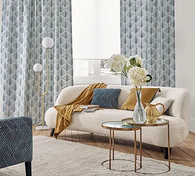 Wohnzimmer mit Holzboden. Links steht ein Sofa in warmem Weisston mit vielen Kissen in ocker und blau. An den Fenstern hängen blaugemusterte Vorhänge. Vor dem Sofa befinden sich zwei kleine Abstelltische mit einer Karaffe und einer Blumenvase. Links im Vordergrund befindet sich ein Sessel.