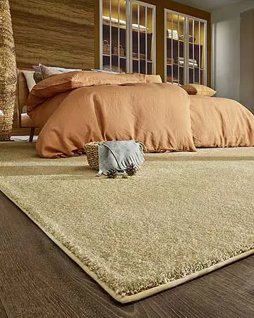 In einem Schlafzimmer liegt ein großer hellbrauner Teppich mit mittelhohem Flor auf einem dunklen Parkettboden. Darauf steht ein Bett mit ockerfarbenen Decken und Kissen. Dahinter ist eine holzvertäfelte Wand mit beleuchteten Vitrinen zu sehen.