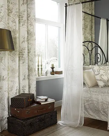 Ein klassisch gemütlich eingerichtetes Schlafzimmer. Die weißen Fenster sind mit hellen, floral gemusterten Vorhängen dekoriert Rechts ist ein Bett mit schwarzen Metallrahmen und einem Baldachin zu sehen, links eine Stehlampe mit bronzefarbenem Schirm und drei alte Lederkoffer.