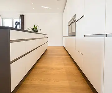 Blick in eine moderne weiße Küche. Links ist ein Küchenblock mit anthrazitfarbener Arbeitsplatte, anthrazitfarbenen Seitenwänden und weißen Schubladen ohne Griffe zu sehen. Rechts sieht man eine vollständig geschlossene Wand aus weißen Küchenschränken, mit integriertem Ofen und komplett ohne Griffe. Der Boden besteht aus einem buchefarbenen Parkett.