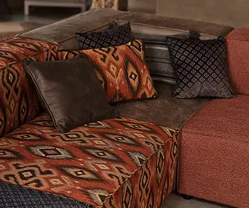 Nahaufnahme eines Ecksofas aus vier Elementen. Jedes Element hat einen anderen Bezug: mit einem dunklen geometrischen Muster, einem afrikanisch anmutenden rot-orangen Muster, braunem Leder und rostbraunem Stoff. Auf dem Sofa liegen 5 Kissen mit den gleichen Bezügen wie die Sofa-Elemente.