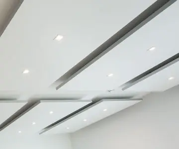 Kassettenförmige, weiße Akustikplatten an einer Decke. Die Platten sind leicht abgehängt und haben einen breiten Abstand zueinander. Die Beleuchtung ist in Form von kleinen Spots integriert.