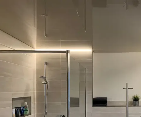 Ein modernes Bad mit einer glänzenden dunkelbraunen Spanndecke. In der Ecke ist eine Dusche mit Glaswand zu sehen, die von der oberen Ecke aus mit einem Lichtband beleuchtet ist.