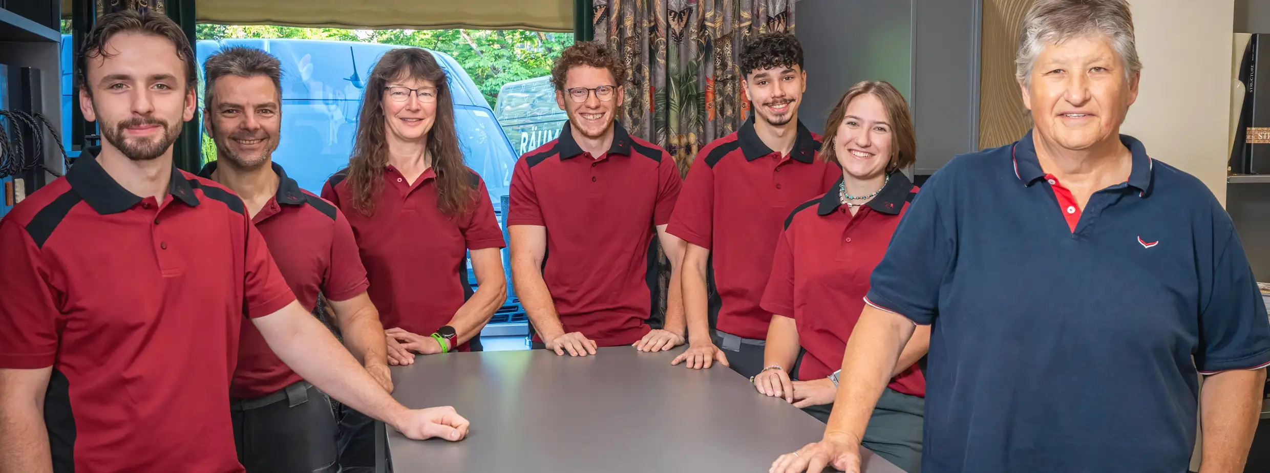 Das Team der Raumausstattung Krebs: 6 Mitarbeiter, davon 2 Frauen, stehen zusammen mit Michael Krebs um einen Arbeitstisch. Die Mitarbeiter sind unterschiedlichen Alters und tragen alle rote Polo-Shirts. Alle lächeln freundlich in die Kamera.