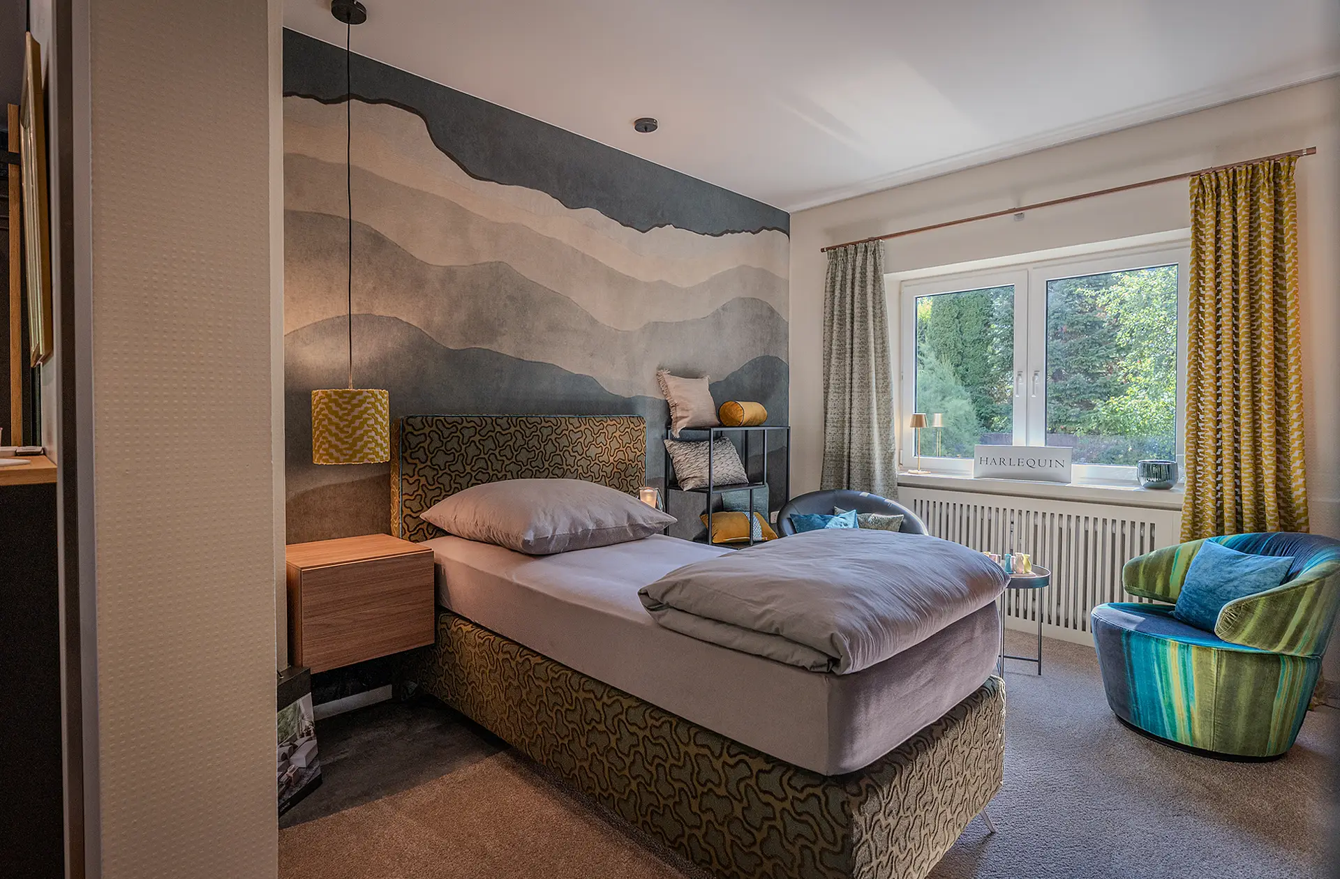 Ein modern eingerichtetes Schlafzimmer mit einem Einzel-Boxspringbett. Das Bett steht in der Mitte. Der untere Teil sowie das Kopfende sind mit einem dunkelbeigen, gemusterten Stoff überzogen. Die Wand hinter dem Bett zeigt eine grafische Tapete mit abgestuften Schattenrissen einer Hügellandschaft. Rechts in der Ecke steht ein moderner bunter Polstersessel in Grün- und Blautönen. Die Vorhänge sind goldfarben und geometrisch gemustert, im gleichen Stoff wie der Lampenschirm der Hängelampe, die neben dem Bett herabhängt.