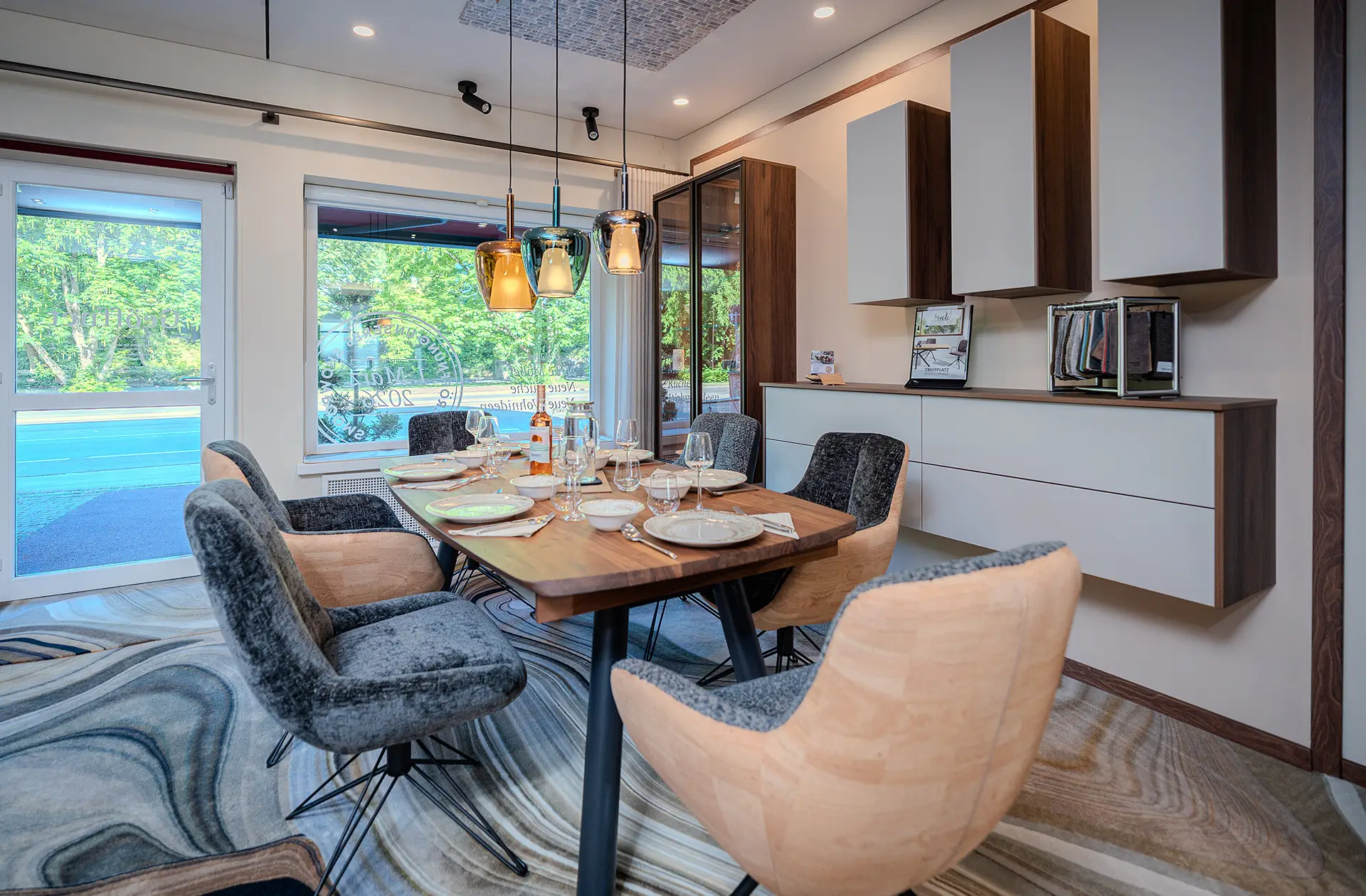 Ausstellungsraum der Raumausstattung Krebs mit einem modernen Esszimmer. In der Mitte steht ein gedeckter Esstisch mit Holzplatte und schwarzen Füßen. Darum 6 mit grauem und beigem Velour gepolsterte, moderne Stühle mit Drahtfüßen. Der Teppich zeigt ein auffälliges Marmor-Muster. Über dem Esstisch hängen 3 Glaslampen, an der Wand daneben sind locker angeordnete Hängeschränke mit weißen Türen und einem Korpus aus dunklem Holz zu sehen.