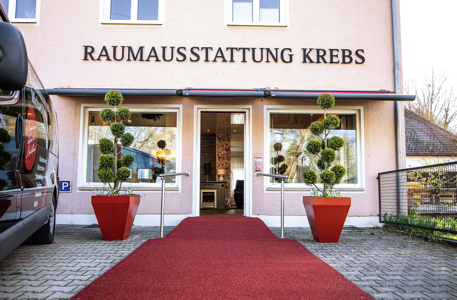 Der Eingangsbereich der Raumausstattung Krebs in Augsburg. Zu sehen sind die Eingangstüre und Schaufenster mit dunkelroten Markisen. 